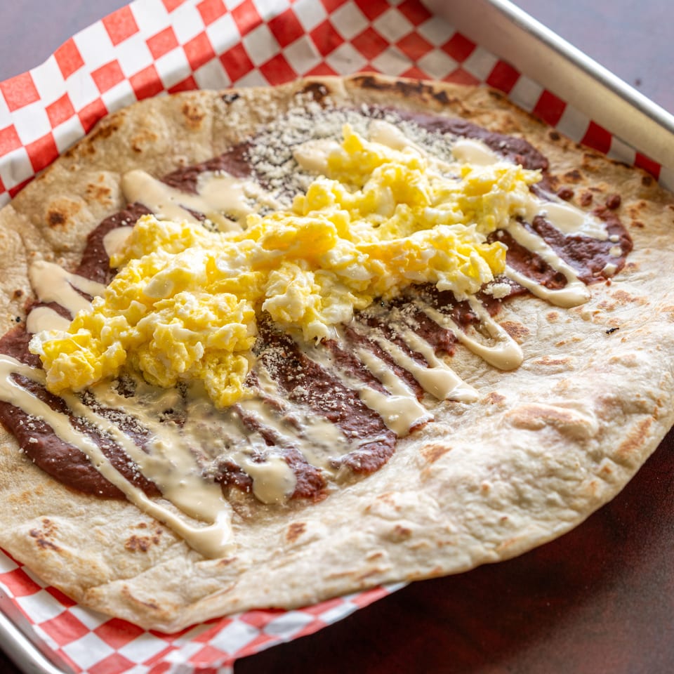 A large flour tortilla topped with refried beans, scrambled eggs, and a drizzle of cream.