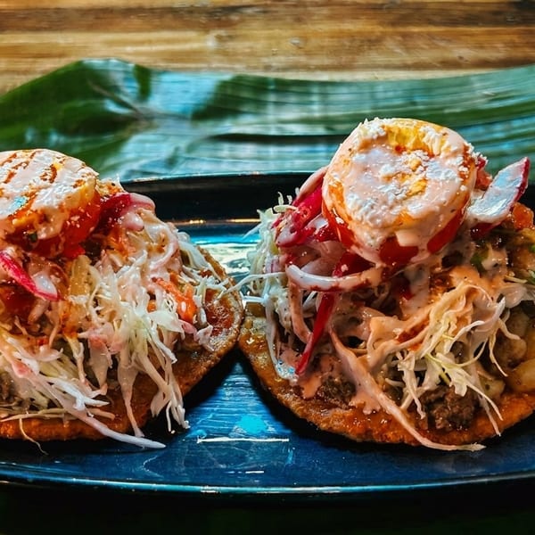 Two vibrant sopes topped with shredded cabbage, meat, sauce, and radishes on a blue plate.