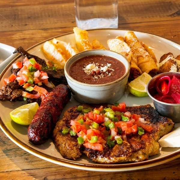 A large platter of grilled meats, sausage, beans, fried plantains, taquitos, and pickled onions.