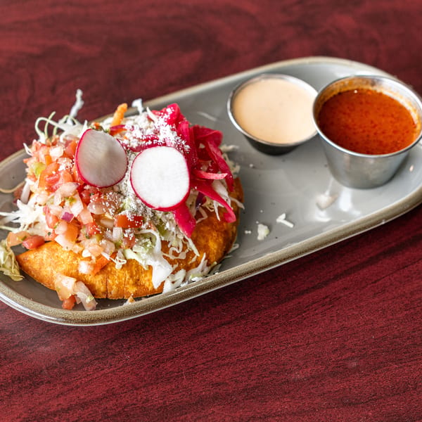 A golden fried empanada topped with pico de gallo, shredded cabbage, radishes, and two dipping sauces.