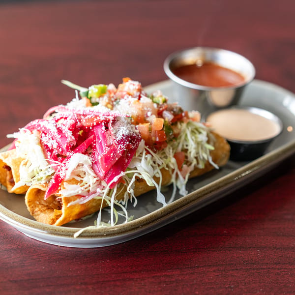 Three golden taquitos topped with shredded cabbage, pickled red onions, pico de gallo, and cheese, with two dipping sauces.