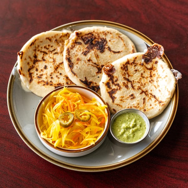 Three pupusas with curtido and green salsa on a plate, on a dark wooden table.