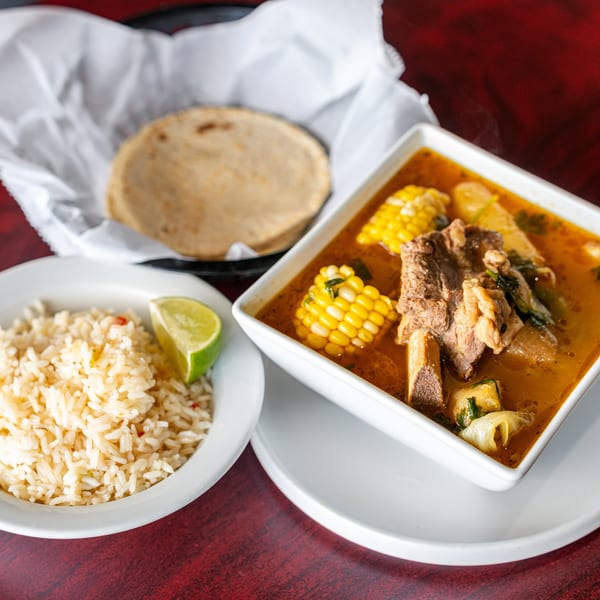 Hearty beef soup with corn and vegetables, served with a side of rice and tortillas.