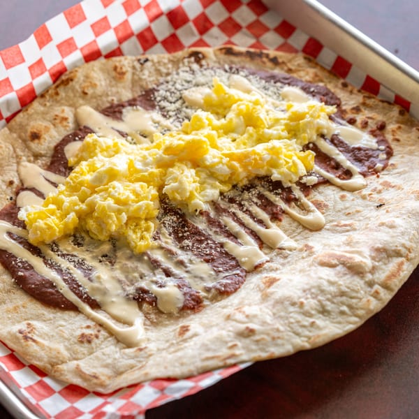 A large flour tortilla topped with refried beans, scrambled eggs, and crema, served in a metal tray.