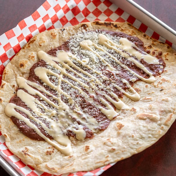 A flatbread topped with refried beans, drizzled with cream, and sprinkled with cheese in a metal tray.